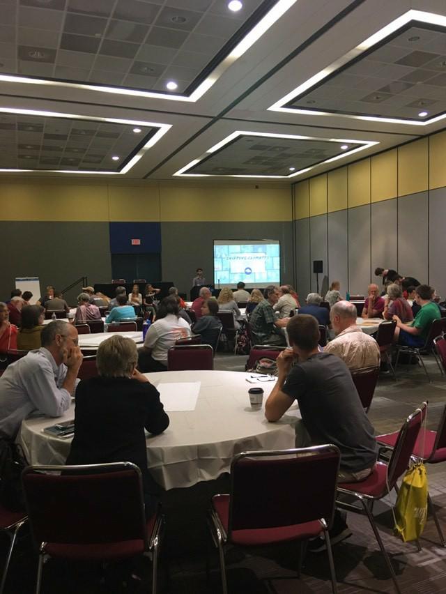 In the distance are the producers of the Shifting Climates podcast giving a presentation. In front of them is a room filled with tables and people around them.