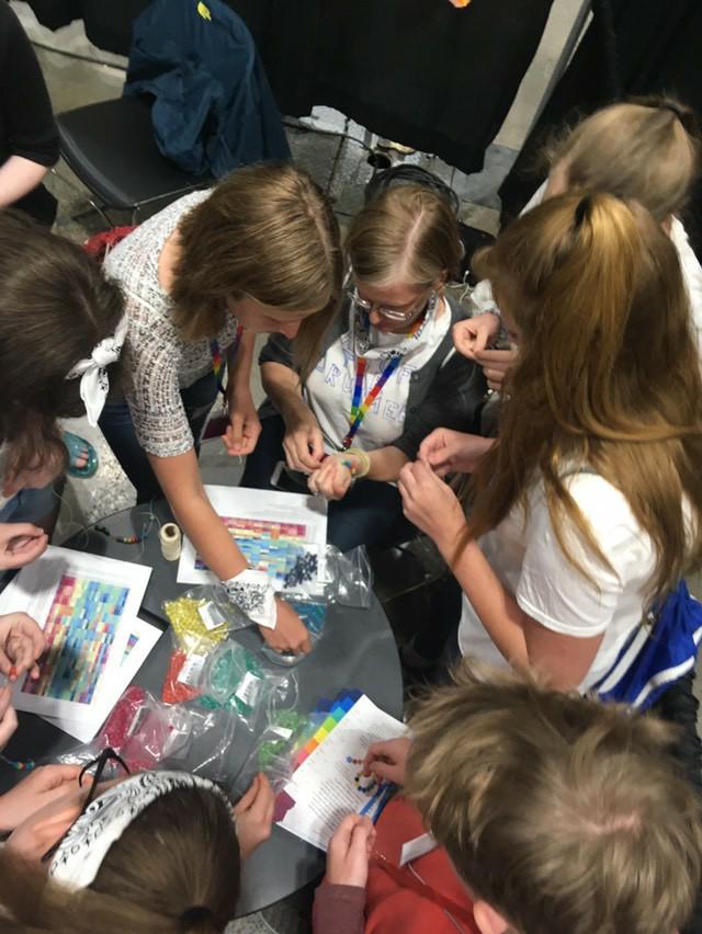 A group of teenagers huddle around a table, on which there are colored charts and matching beads, which they are using to make temperature bracelets.