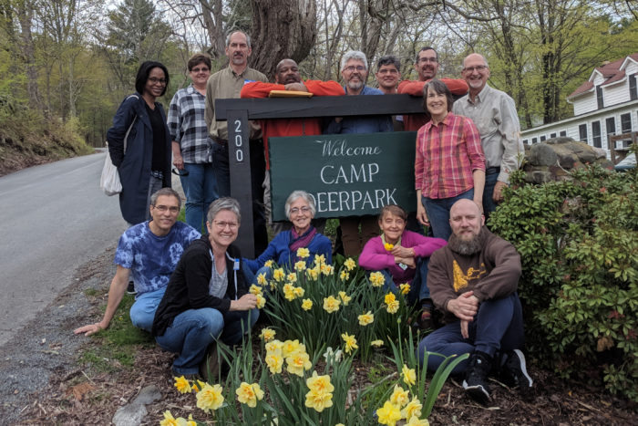 A group of people cluster around a sign reading, 'Welcome Camp Deerpark.' In front of them are blooming daffodils.